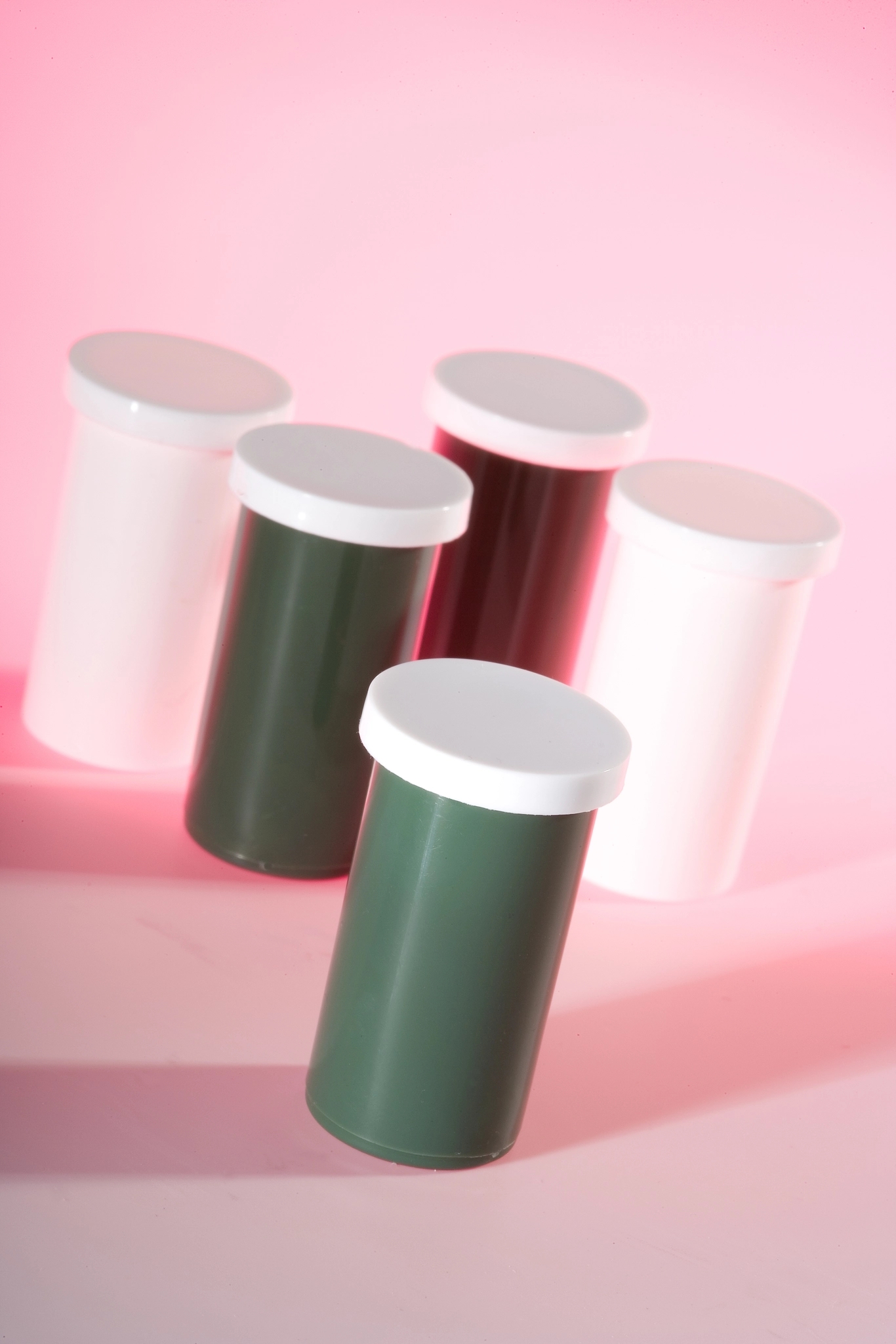 Five cylindrical containers with white caps on a pink background. Two white, two dark green, one dark red.