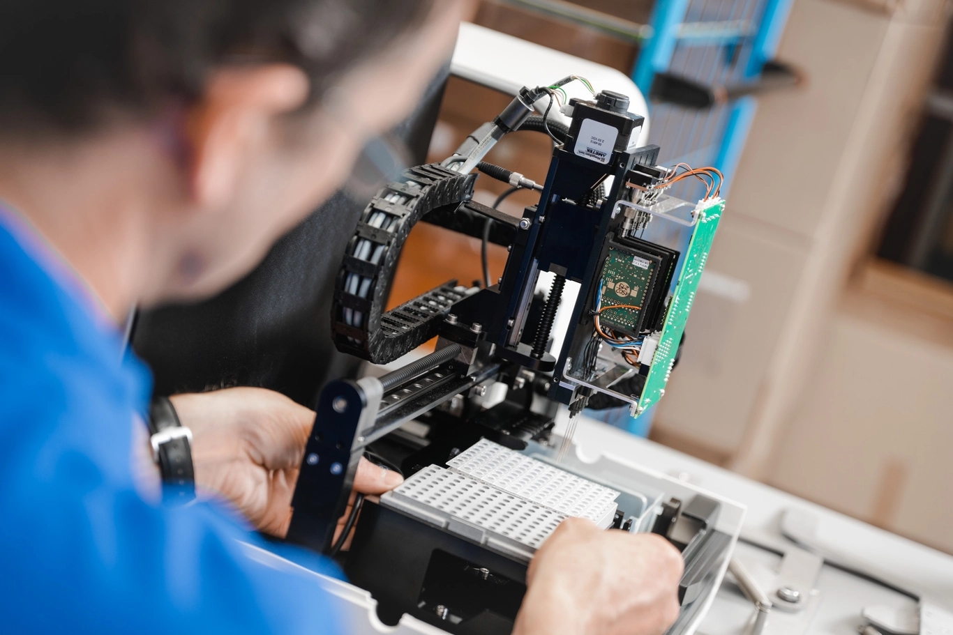 Person in blue shirt repairing a complex automated machine, with exposed circuit boards and mechanical tracks.