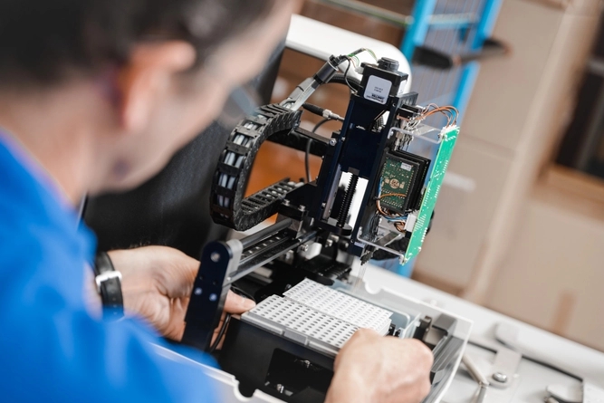 Person in blue shirt repairing a complex automated machine, with exposed circuit boards and mechanical tracks.