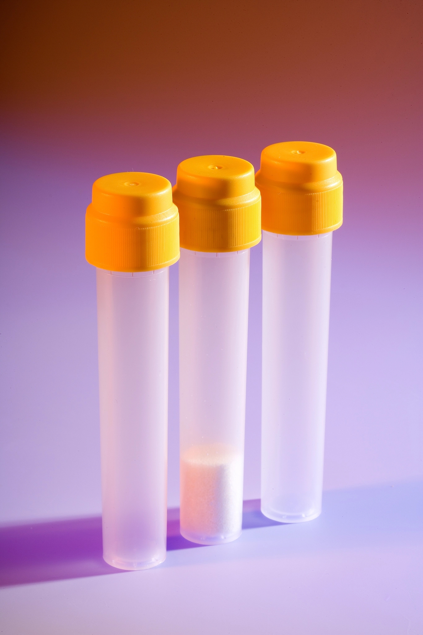 Three frosted tubes with yellow caps standing upright on a purple and orange gradient background. Middle tube has white powder.